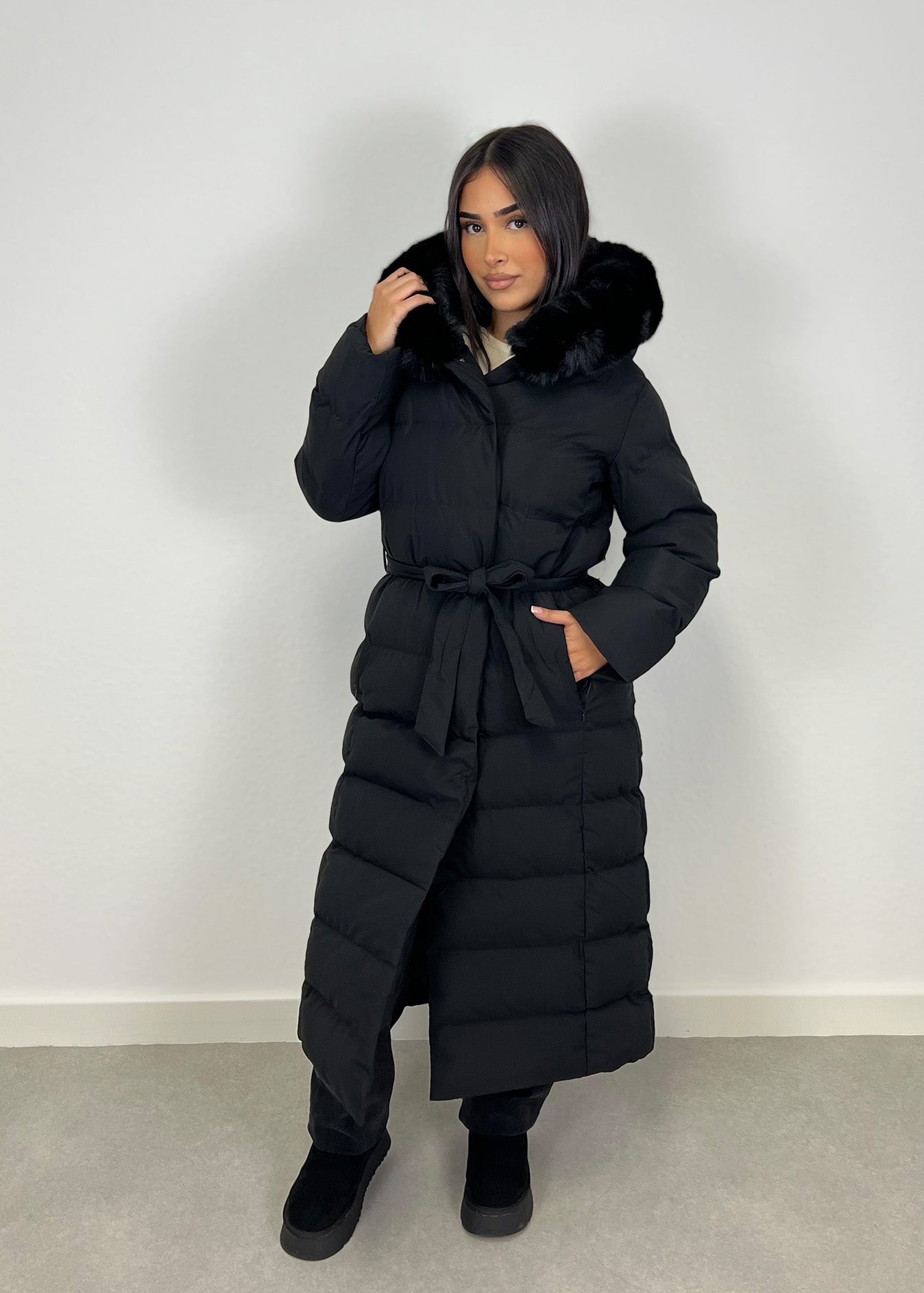 Modell trägt einen langen Damen Steppmantel mit Kapuze und Taillengürtel – NAHA Wintermode