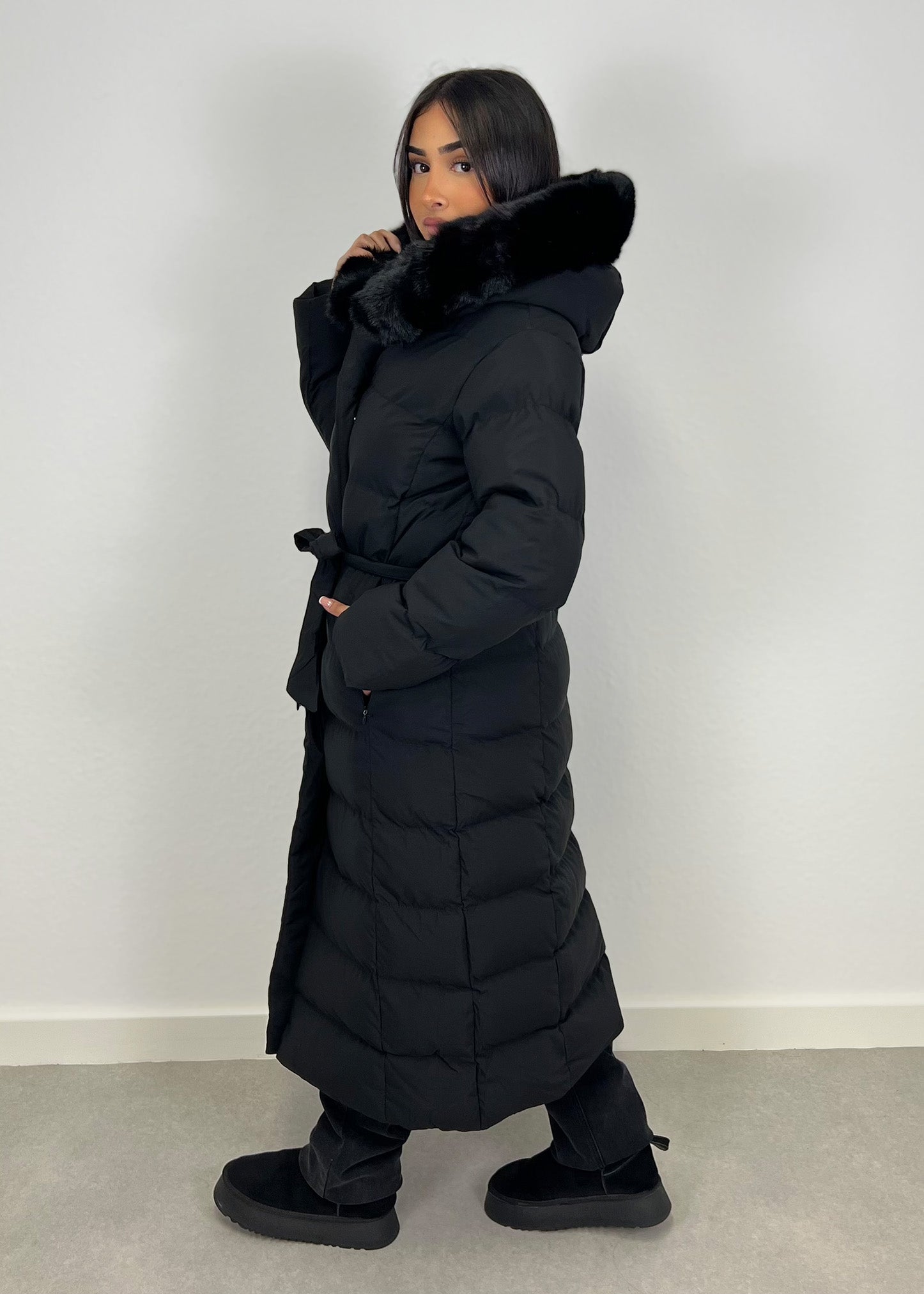 Modell trägt einen langen Damen Steppmantel mit Kapuze und Taillengürtel – NAHA Wintermode