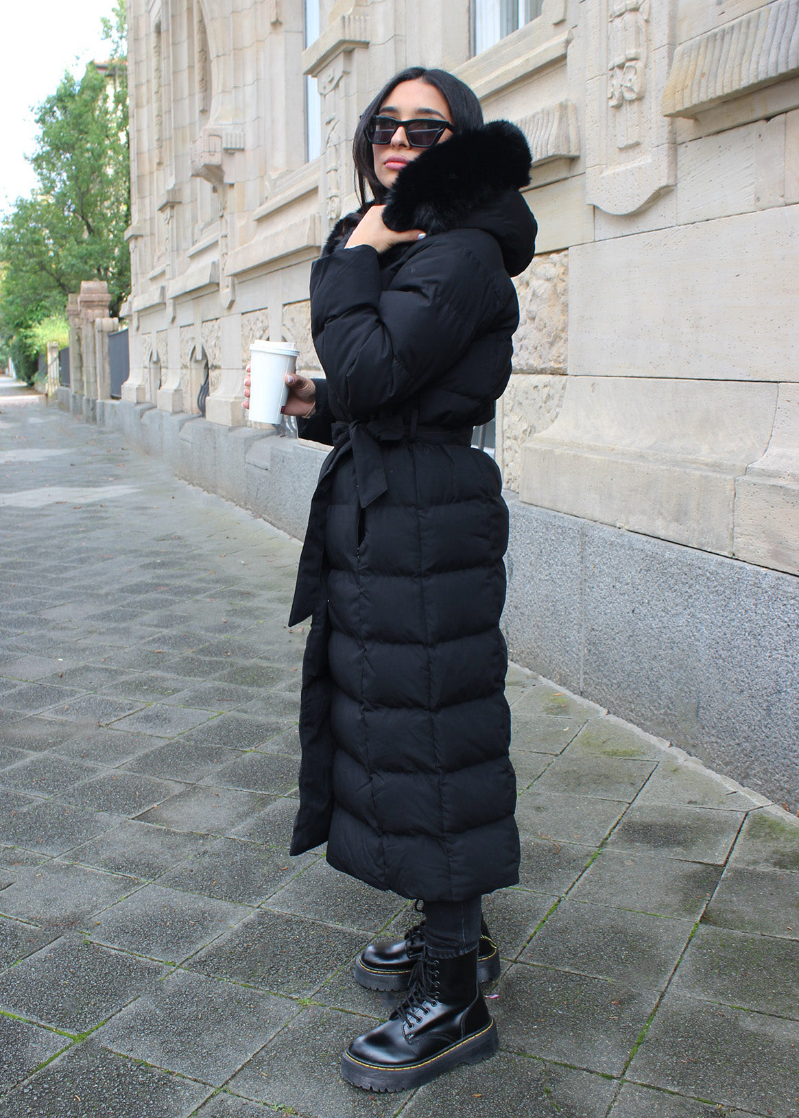 Modell trägt einen langen Damen Steppmantel mit Kapuze und Taillengürtel – NAHA Wintermode