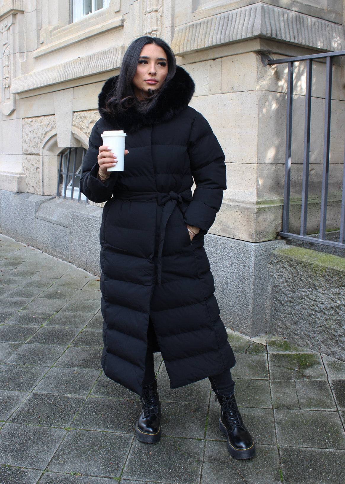 Modell trägt einen langen Damen Steppmantel mit Kapuze und Taillengürtel – NAHA Wintermode