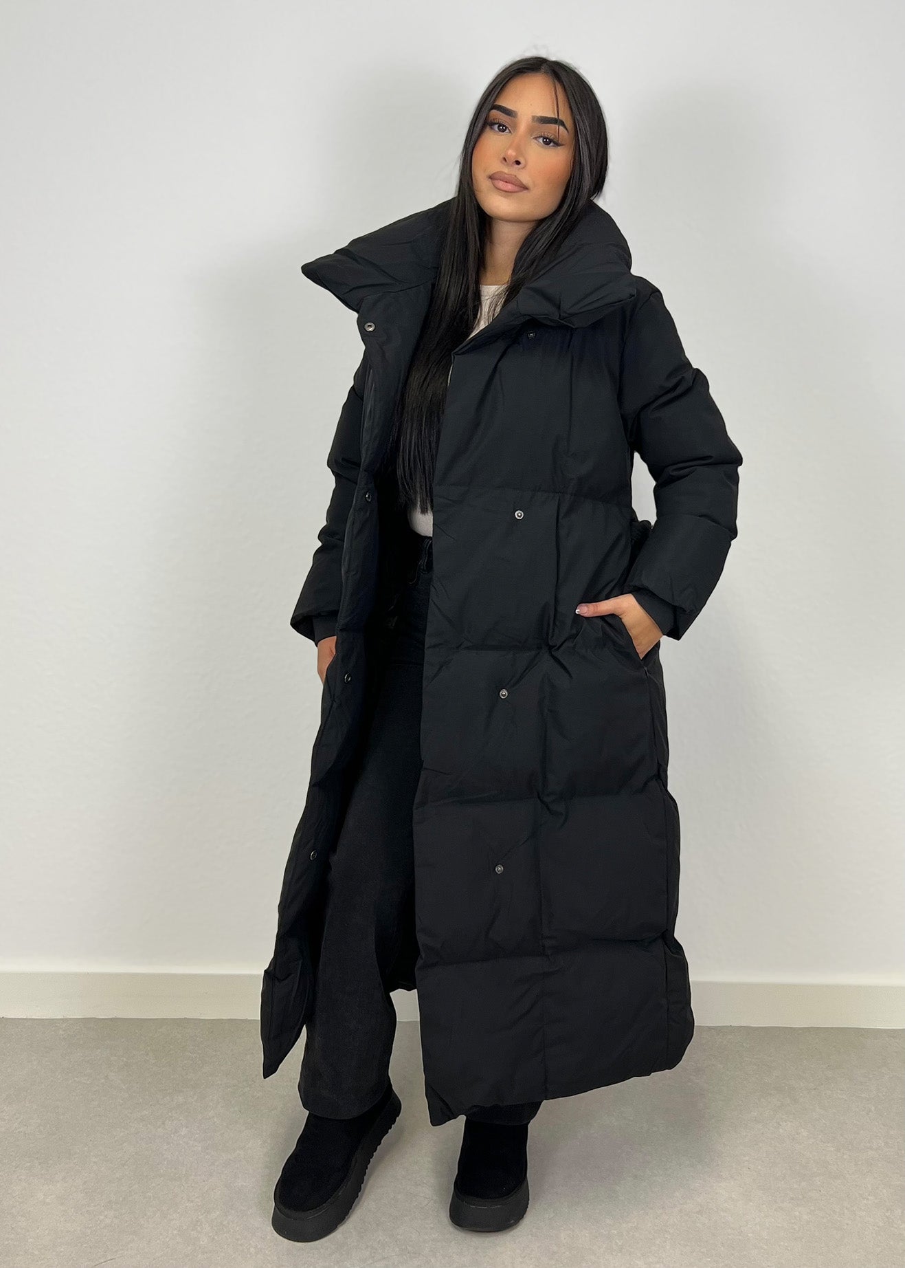Frau trägt lange Winterjacke in Schwarz mit Gürtel & hohem Kragen – eleganter Winterlook von NAHA