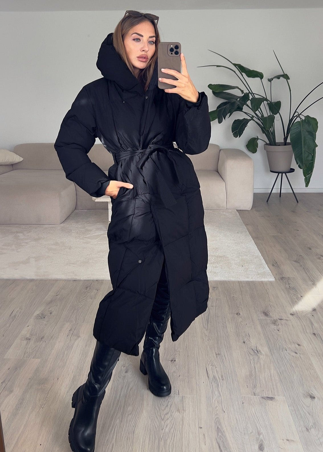 Modell trägt langen Damen Steppmantel in Schwarz mit Kapuze und Taillengürtel – NAHA Winterjacke