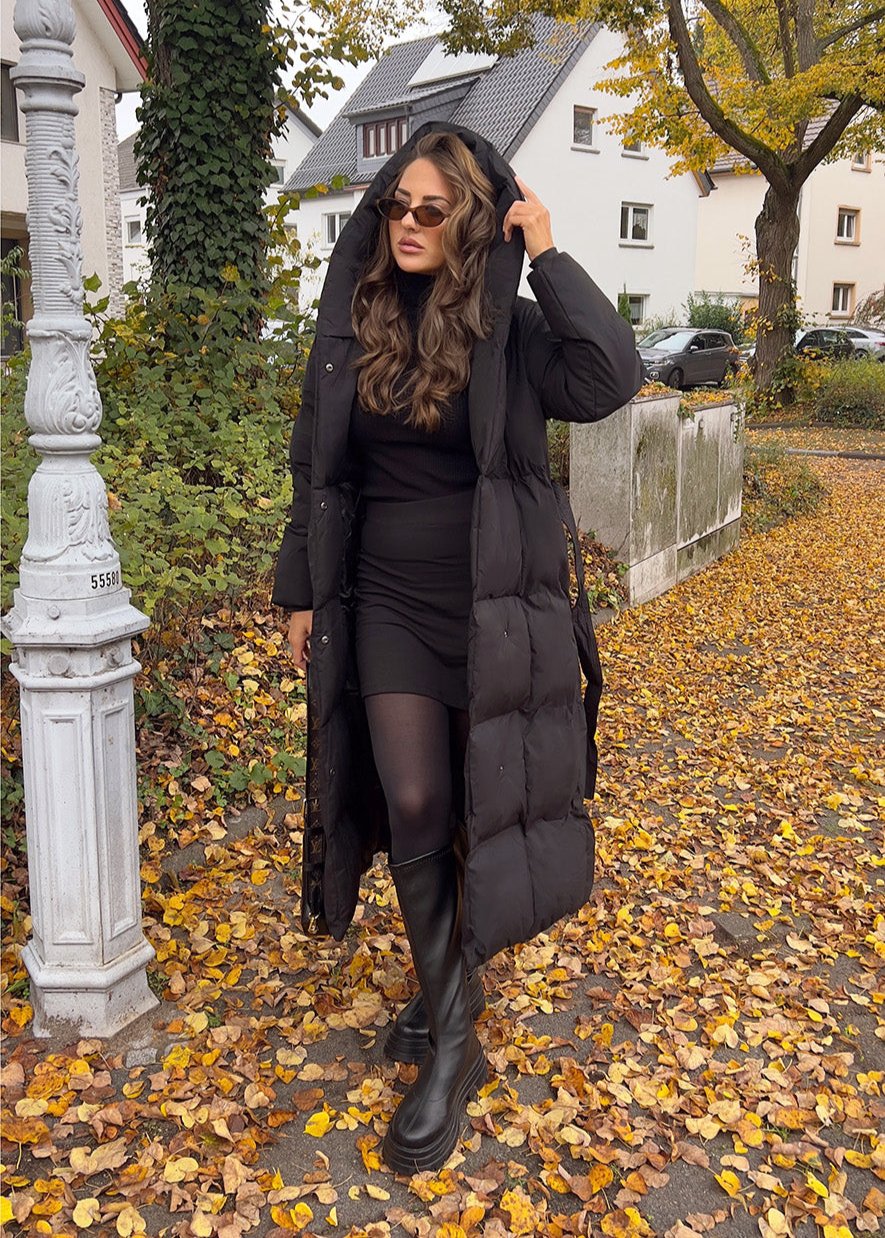 Modell trägt langen schwarzen Damen Steppmantel mit Kapuze und Taillengürtel, kombiniert mit Kleid und Stiefeln im herbstlichen Streetstyle – NAHA Winterjacke
