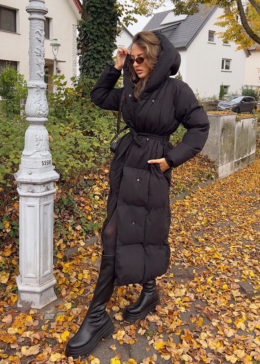 Modell trägt langen schwarzen Damen Steppmantel mit Kapuze und Taillengürtel, herbstlicher Outdoor-Look mit Chunky Boots – NAHA Winterjacke