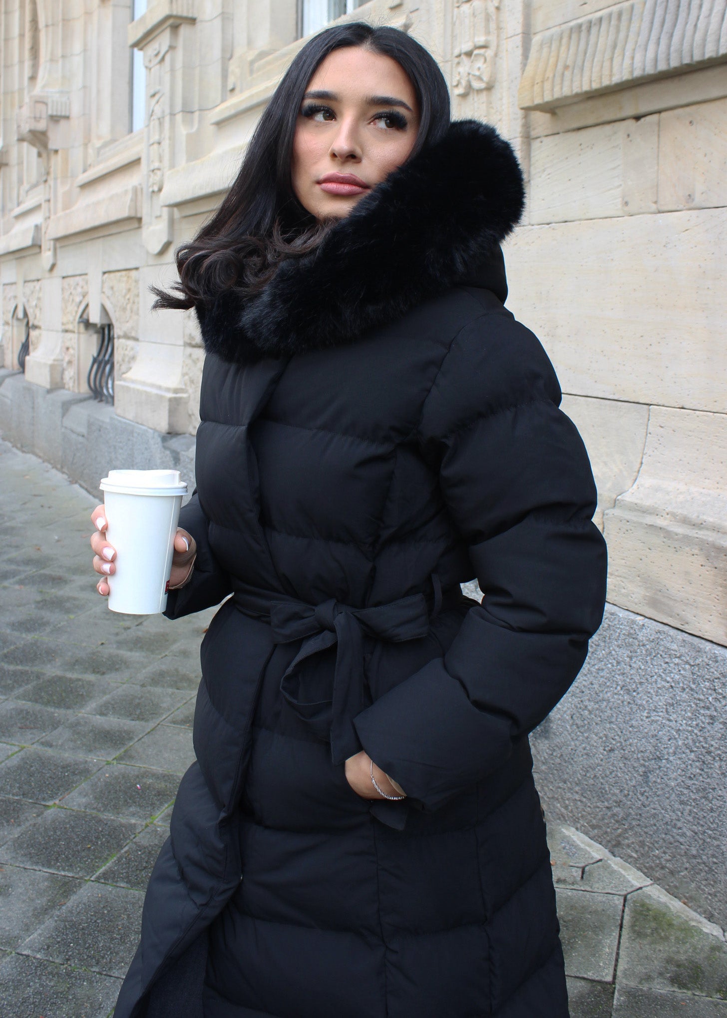 Frau trägt eine lange schwarze Winterjacke mit Gürtel und Fellkapuze – eleganter Look für kalte Tage
