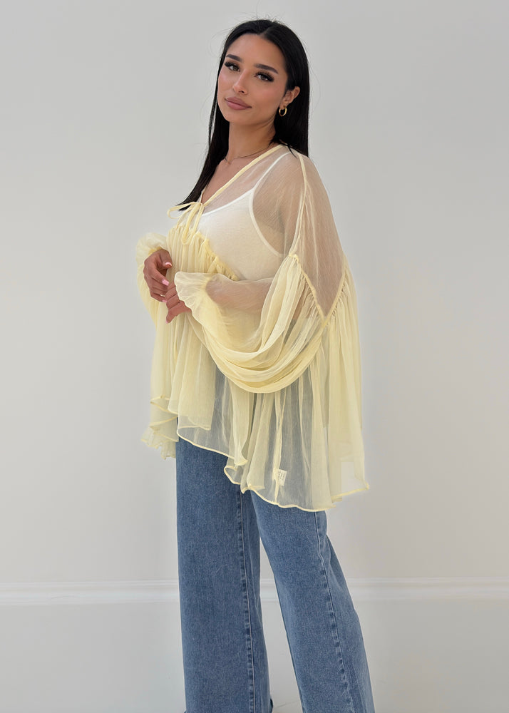 Frau trägt transparente Rüschenbluse mit weitem Schnitt, Volantärmeln & Bindeband – eleganter Sommerlook von NAHA