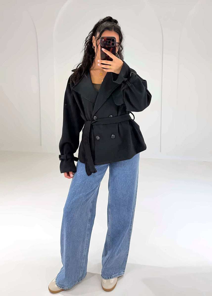 Frau in schwarzem Cropped Trenchcoat mit Bindegürtel zu Wide-Leg-Jeans, ganze Figur