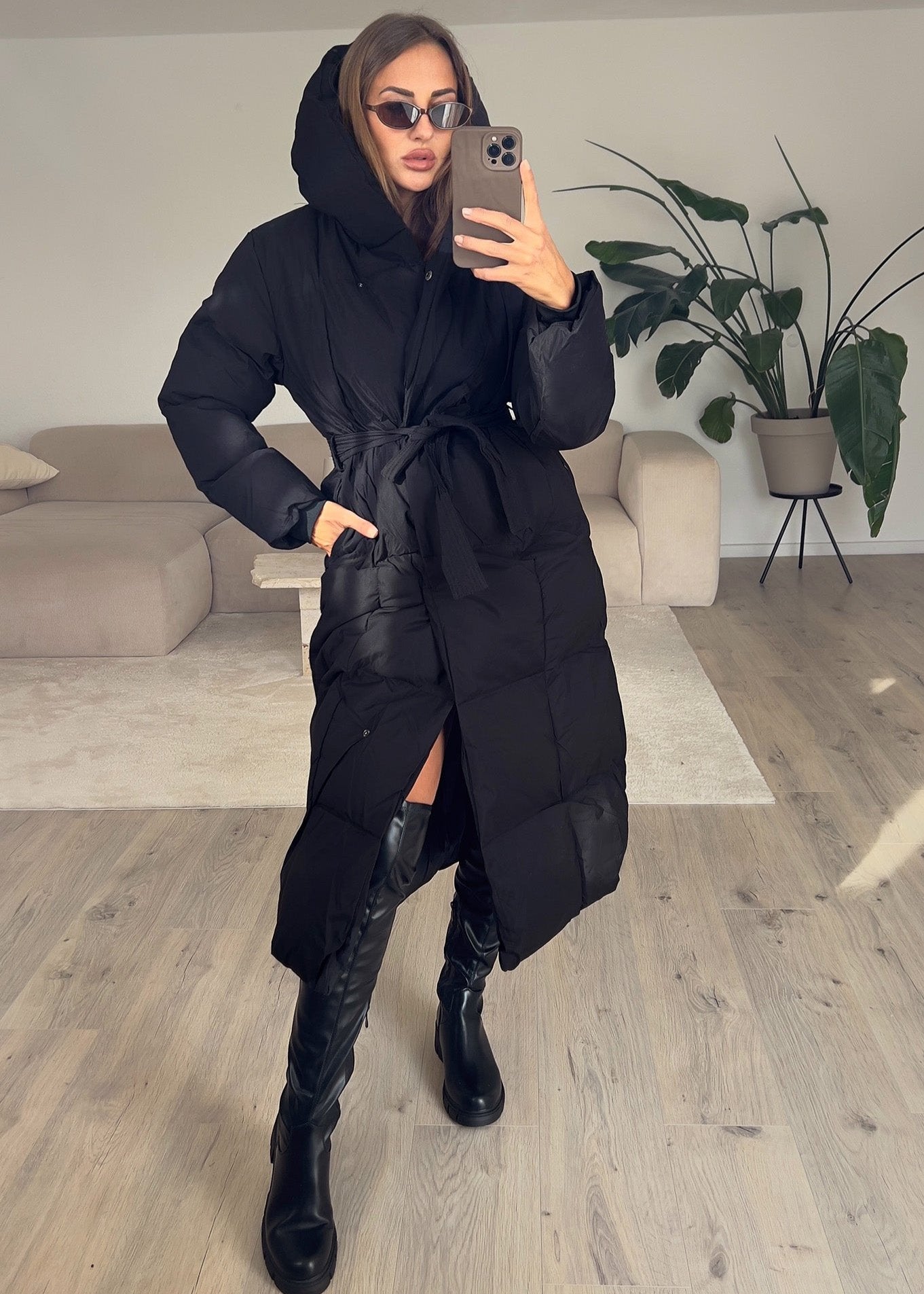 Modell trägt langen schwarzen Damen Steppmantel mit Kapuze und Taillengürtel, geschlossen – NAHA Jacke