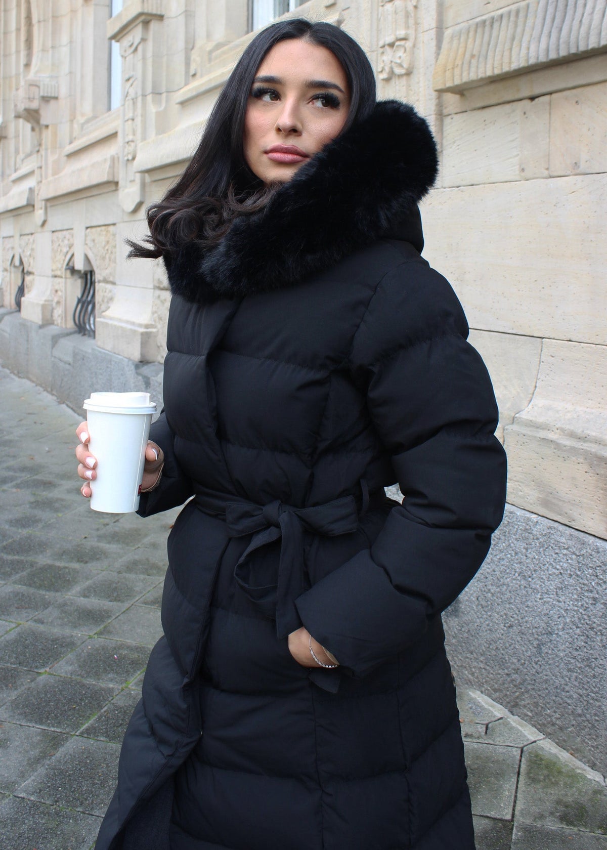 Modell trägt einen langen Damen Steppmantel mit Kapuze und Taillengürtel – NAHA Wintermode