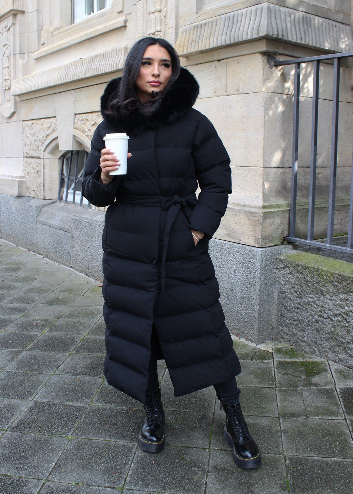 Modell trägt einen langen Damen Steppmantel mit Kapuze und Taillengürtel – NAHA Wintermode