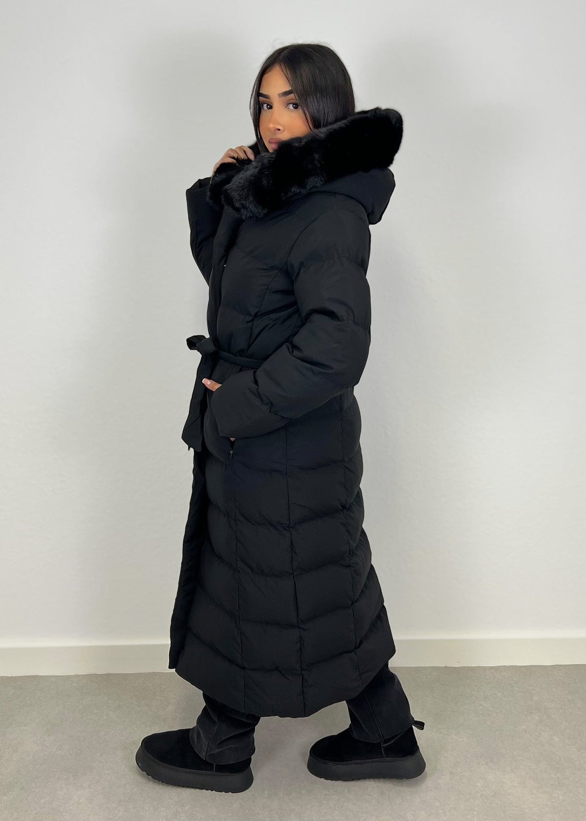 Modell trägt einen langen Damen Steppmantel mit Kapuze und Taillengürtel – NAHA Wintermode