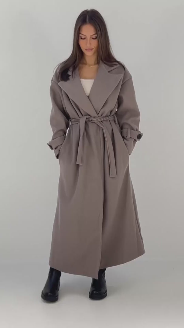 Langer Damen Oversize-Mantel in Taupe mit breitem Revers und Bindegürtel, kombiniert mit schwarzen Boots – NAHA