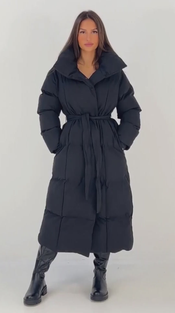 Lange schwarze Winterjacke für Damen: Puffermantel mit hohem Stehkragen und Bindegürtel, getragen mit schwarzen Stiefeln – NAHA