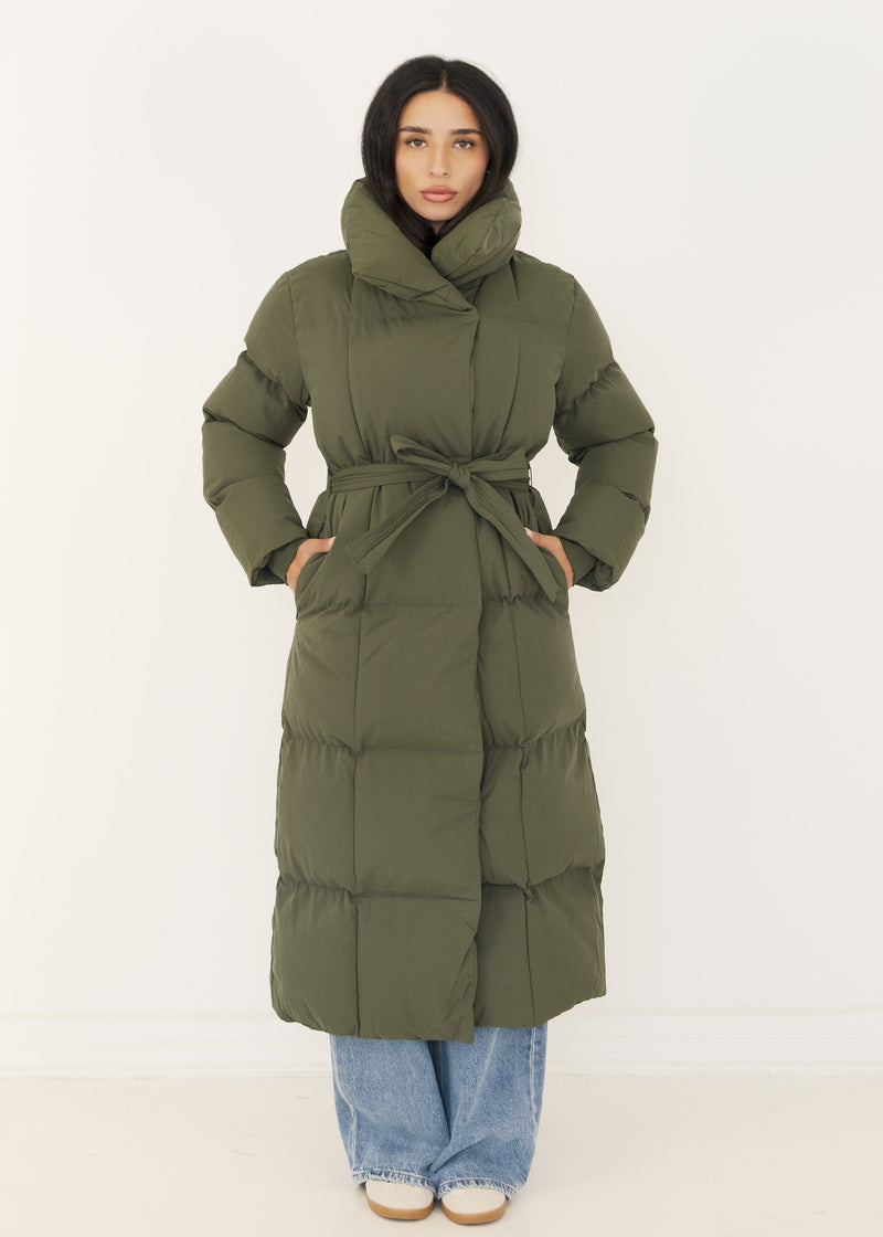 Frau in langem khakifarbenem Puffer Mantel mit Gürtel, Hände in den Taschen